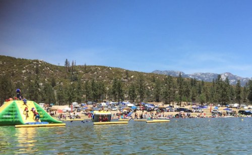 A sunny lakeside scene with people enjoying inflatable slides and floating platforms, surrounded by trees and mountains.