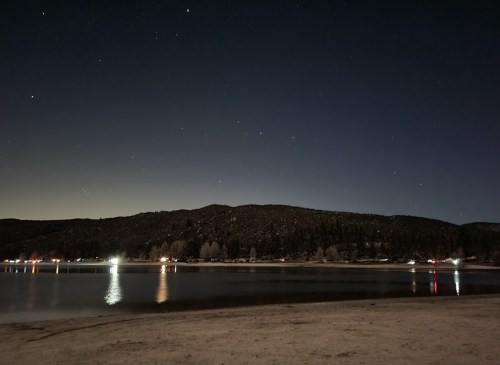 A serene night scene of a lake with a dark hillside and stars above, reflecting lights from nearby buildings.