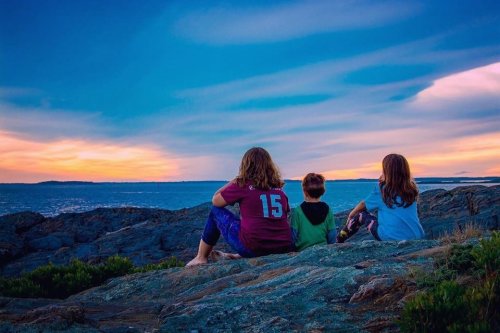 Three children sit on a rocky shore, watching a colorful sunset over the ocean.