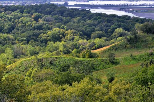 Lush green hills and trees stretch across the landscape, with a river visible in the distance under a clear sky.
