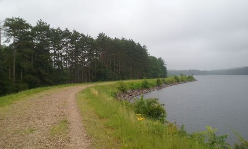 A gravel path curves along a calm lake, bordered by lush greenery and trees under a cloudy sky.