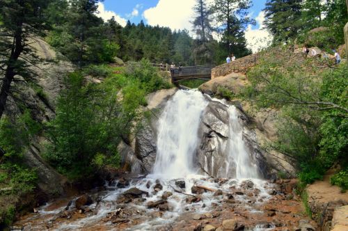 A scenic waterfall cascades over rocks, surrounded by lush greenery and a bridge in the background.