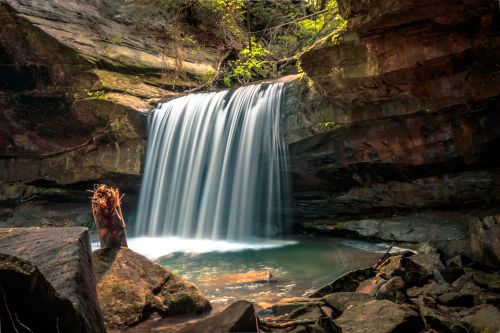 A serene waterfall cascades over rocks into a tranquil pool, surrounded by lush greenery and natural stone formations.
