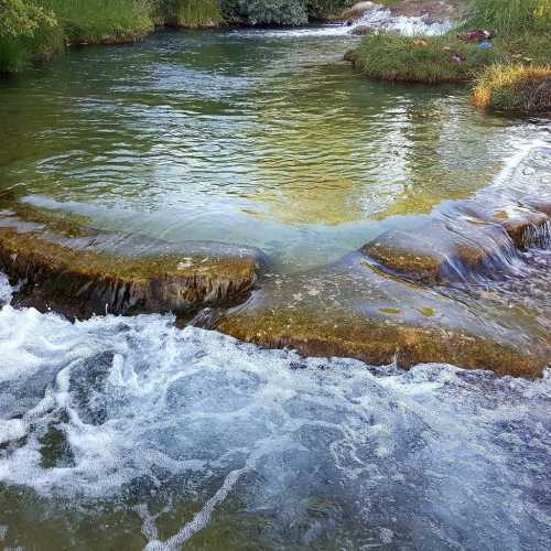 A serene river scene with clear water flowing over rocky steps, surrounded by lush greenery and gentle ripples.