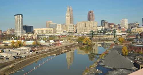 City skyline with tall buildings reflecting in a river, surrounded by autumn trees and industrial areas. Clear blue sky.