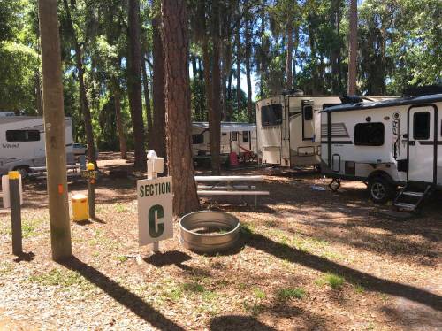 A campsite with several RVs parked among tall trees, featuring a sign for "Section C" and a picnic table nearby.