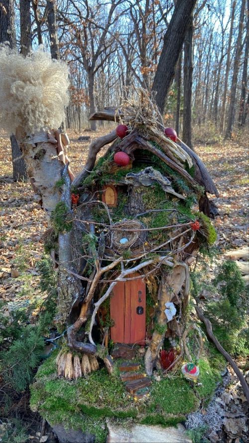 A whimsical fairy house made of natural materials, nestled among trees in a forest setting, adorned with moss and decorations.