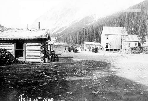 Historic black-and-white photo of a small settlement with log cabins and a group of people in a mountainous landscape.