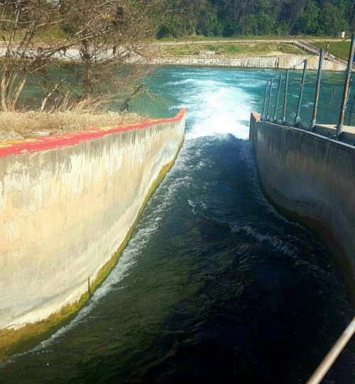 A narrow water channel with flowing water, flanked by concrete walls and greenery in the background.