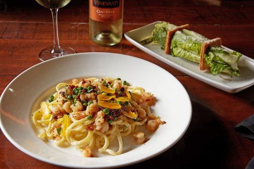 A plate of pasta with shrimp and peas, alongside a Caesar salad and a glass of white wine on a wooden table.