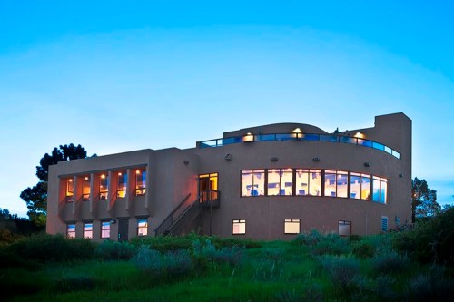 A modern adobe-style building with large windows, surrounded by greenery, under a twilight sky.