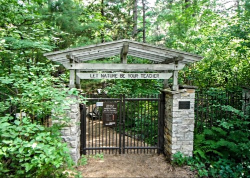 A wooden gate surrounded by greenery, with a sign that reads "LET NATURE BE YOUR TEACHER."