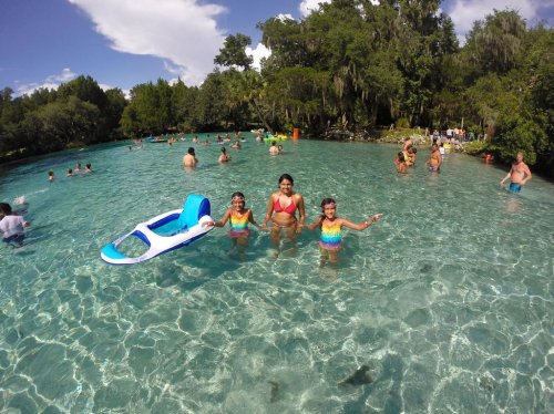 Three children in colorful swimsuits play in a clear, blue swimming area, surrounded by other people enjoying the water.