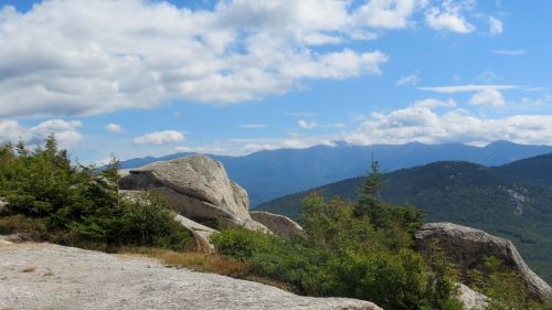 A scenic view of mountains under a blue sky with clouds, featuring large rocks and greenery in the foreground.