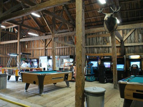 A rustic barn interior with pool tables, arcade games, and a mounted deer head on the wall.