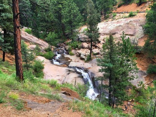 A serene landscape featuring a small waterfall cascading over rocks, surrounded by lush green trees and a rocky terrain.