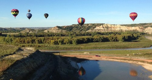 Colorful hot air balloons float over a scenic landscape with hills, trees, and a winding river under a clear blue sky.