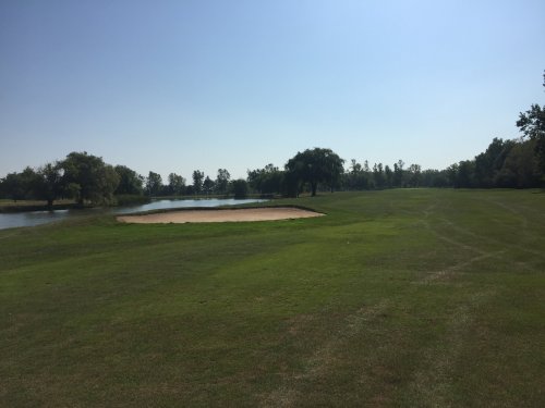A sunny golf course scene featuring a sand trap, lush green grass, and a calm pond surrounded by trees.