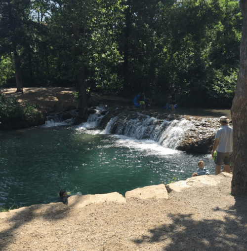 A serene natural scene featuring a small waterfall and people enjoying the water in a lush, green park.