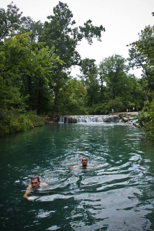 Two people swim in a clear, greenish-blue river near a small waterfall, surrounded by lush trees.