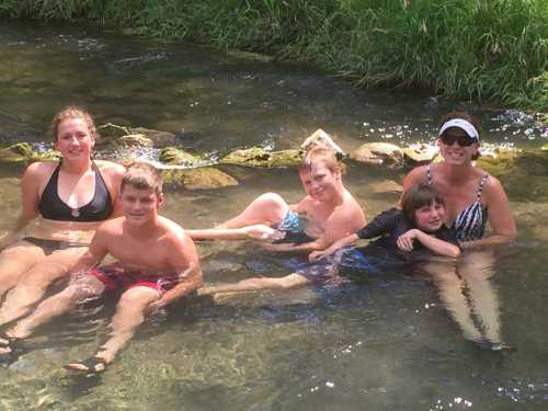 A group of five people, including children and an adult, enjoying a sunny day in a shallow stream.