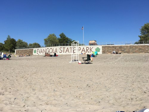 A sandy beach at Evangola State Park with a large sign and people relaxing in the background under a clear blue sky.