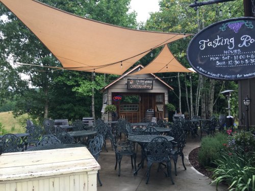 Outdoor seating area with metal tables and chairs, shaded by large fabric canopies, near a tasting room with an open sign.
