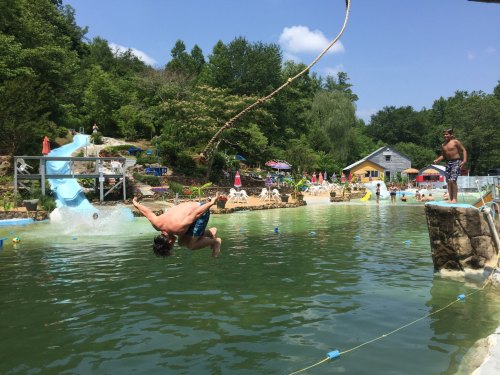 A person jumps off a rock into a swimming area, with slides and people enjoying the water in the background.