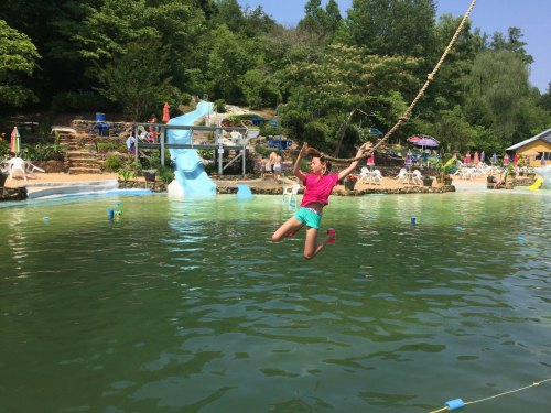 A girl swings over a green lake, preparing to jump into the water, with a water slide and trees in the background.