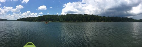 A panoramic view of a calm lake surrounded by lush green trees under a partly cloudy sky.