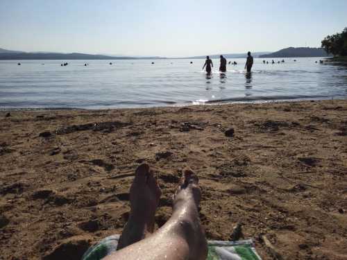 A sandy beach with a person’s feet in the foreground and people swimming in the water in the background.