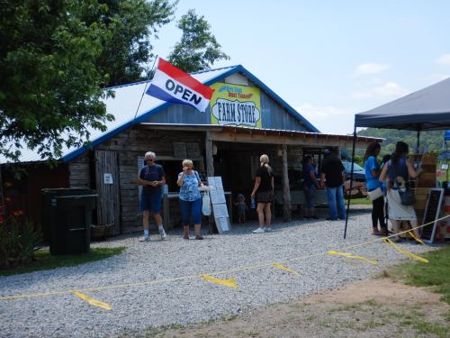 A rustic farm store with an "Open" sign, people browsing outside, and a tent nearby on a sunny day.