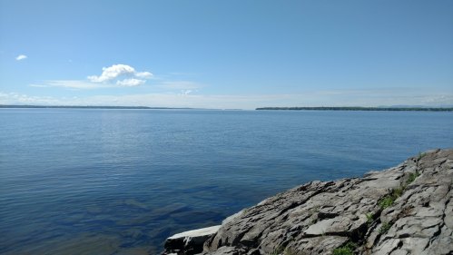 A calm lake under a clear blue sky, with rocky shoreline and distant land on the horizon.