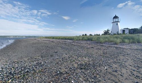 A sandy beach with pebbles and grass, featuring a white lighthouse against a clear blue sky.