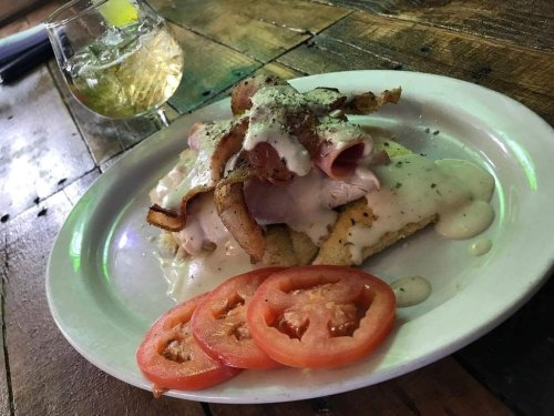A plate of food featuring sliced tomatoes, bacon, ham, and creamy sauce, with a glass of drink in the background.
