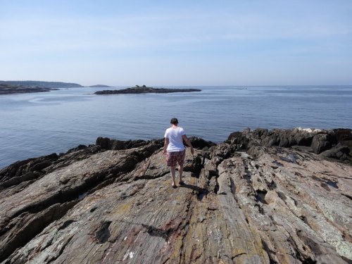 A person walks along rocky shorelines, with calm waters and distant islands under a clear blue sky.