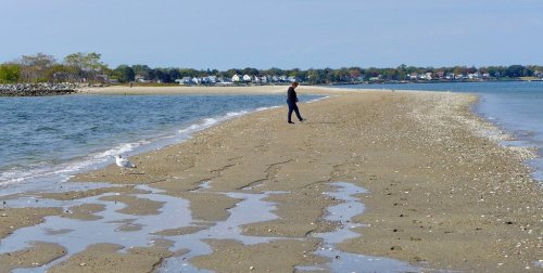 A person walks along a sandy shore with water on both sides, surrounded by a calm landscape and distant houses.