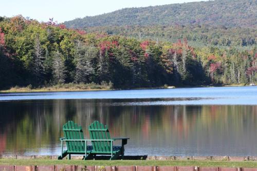 Two green Adirondack chairs by a calm lake, surrounded by trees with autumn foliage reflecting in the water.