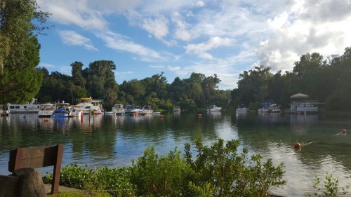 A serene waterway with boats anchored, surrounded by lush greenery and a partly cloudy sky.