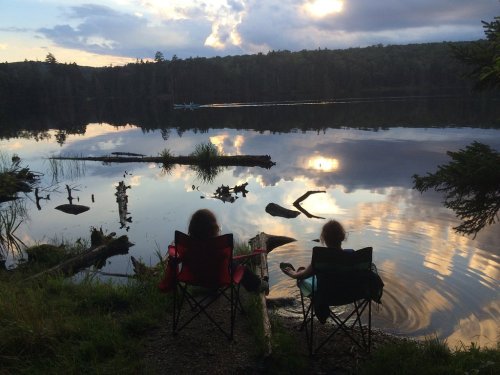 Two people sit in chairs by a calm lake, reflecting trees and clouds, with a boat in the distance under a sunset sky.