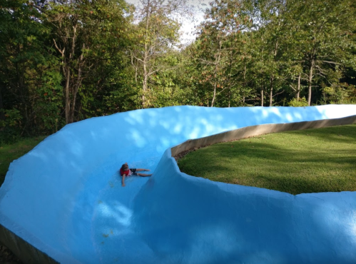 A child slides down a blue water slide surrounded by green trees and grass.