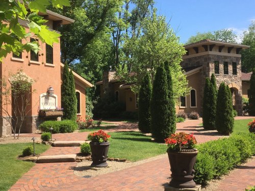 A landscaped garden featuring stone and stucco buildings, brick pathways, and potted plants under a clear blue sky.