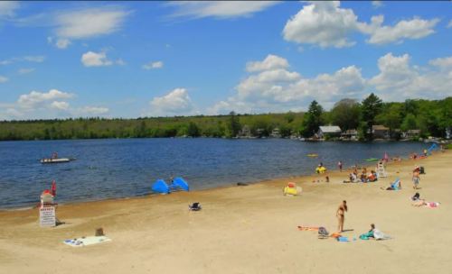 A sandy beach by a lake with people sunbathing, swimming, and enjoying a sunny day under a blue sky with clouds.