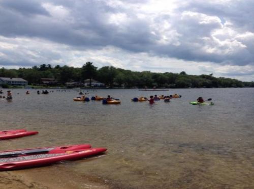 A group of people kayaking on a lake with cloudy skies, while others swim nearby and red kayaks rest on the shore.
