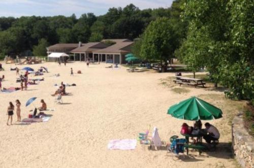 A sandy beach scene with people sunbathing, umbrellas, and a building in the background surrounded by trees.