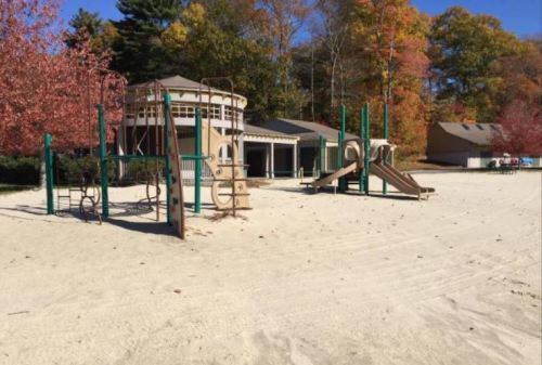 Playground with slides and climbing structures on sandy ground, surrounded by trees with autumn foliage.