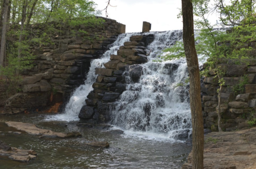 A cascading waterfall flows over stone steps, surrounded by lush greenery and a calm river below.
