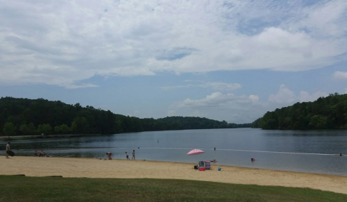 A serene lake scene with a sandy beach, people enjoying the water, and a pink umbrella under a partly cloudy sky.