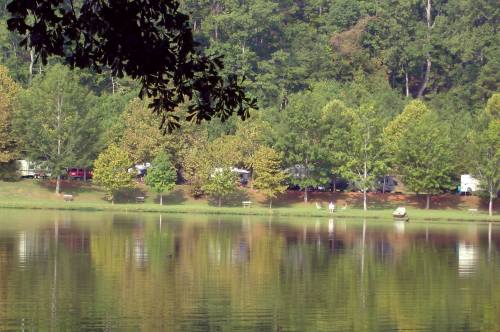 A serene lake reflecting trees and campers along the shore, surrounded by lush greenery.