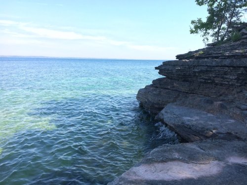 A rocky shoreline meets a calm, clear blue lake under a bright sky. Greenish water gently laps against the stones.